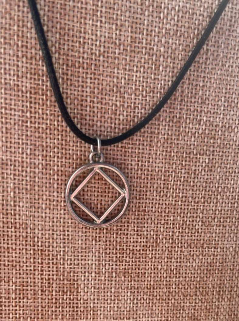 3/4 inch alloy NA symbol charm affixed to a black cord necklace displayed on a tan jewelry holder with the sunlight facing the camera