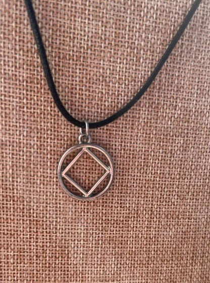 3/4 inch alloy NA symbol charm affixed to a black cord necklace displayed on a tan jewelry holder with the sunlight facing the camera