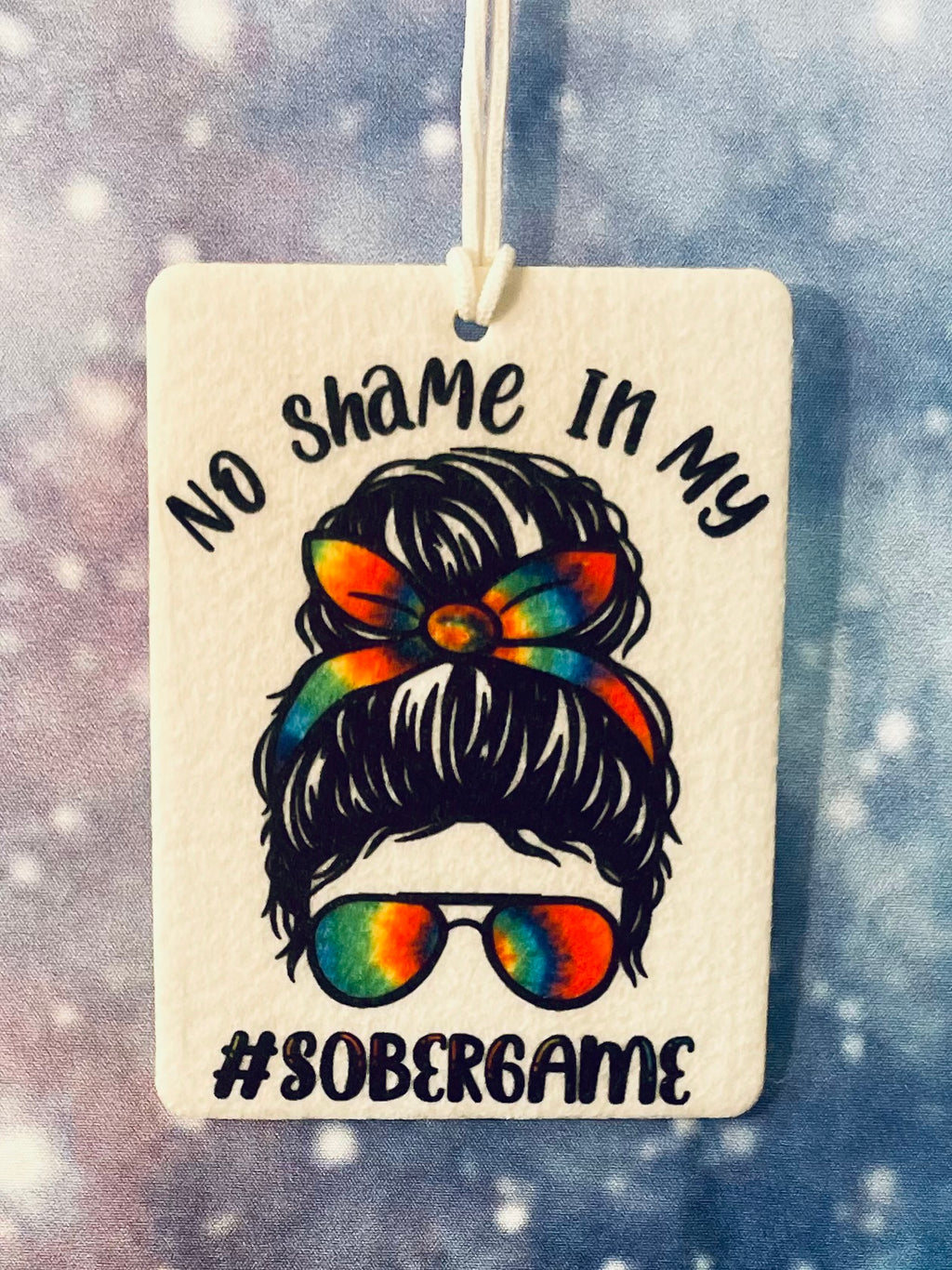 Decorative tag with a design of a woman's head with a bun and sunglasses, text 'No Shame In My #SoberGame', against a snowy background.